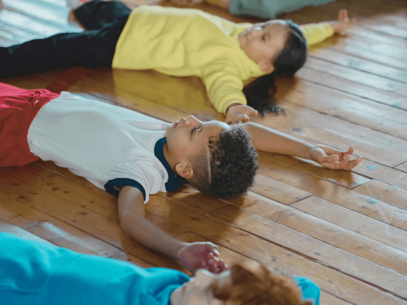 drei Kinder liegen auf einem Holzboden nebeneinander und strecken die Arme bei einer Aktivierungsübung
