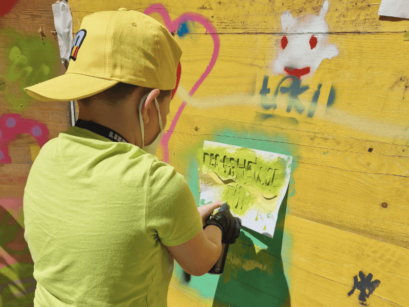 ein Grundschulkind mit einem gelben Cap sprüht mit einer Schablone Graffiti an die Wand