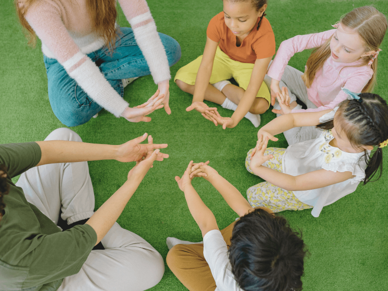 vier Kinder sitzen mit zwei Erzieherinnen im Kreis und strecken ihre Arme mit verschränkten Fingern nach vorn