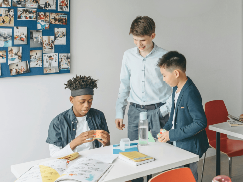 zwei Schüler stehen um einen Schreibtisch, an dem ein anderer Schüler sitzt