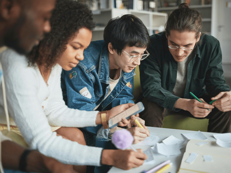 vier Schüler*innen beugen sich über einen Tisch auf dem sie Vokabeln auf Karteikarten schreiben und ihren DaZ-Wortschatz üben